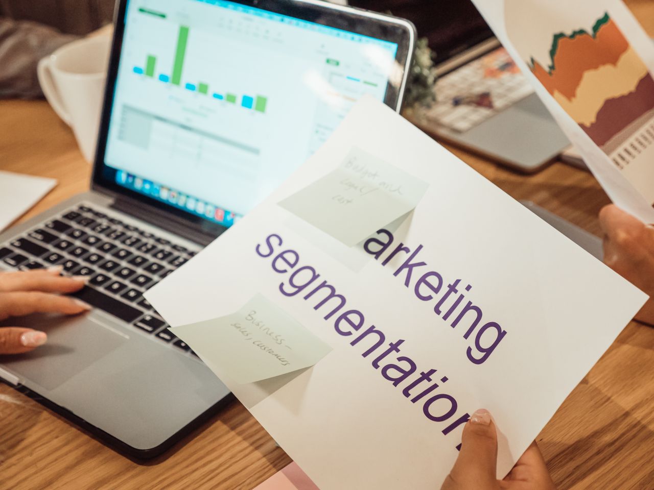 A contemporary work environment showing hands reviewing marketing data on a laptop and printed documents.