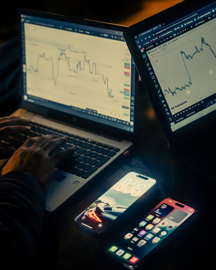 about-us-01 A trading workspace with laptops displaying charts and smartphones, set in a low light environment, Dhaka.